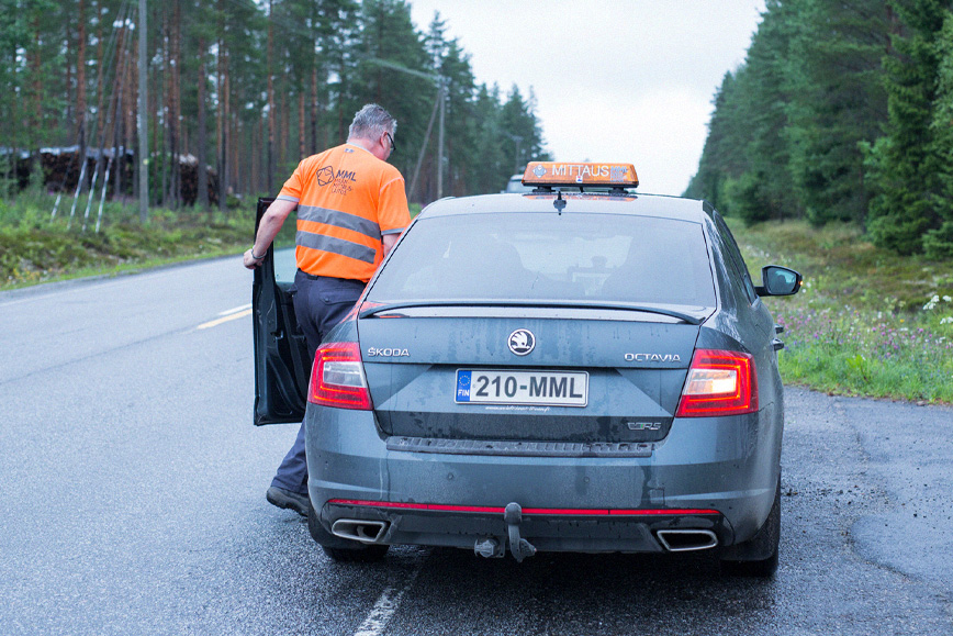 Oranssipaitainen Maanmittauslaitoksen kartoittaja on astumassa autoon. Auto on pysäköity asfalttitien levikkeelle metsän reunaan.