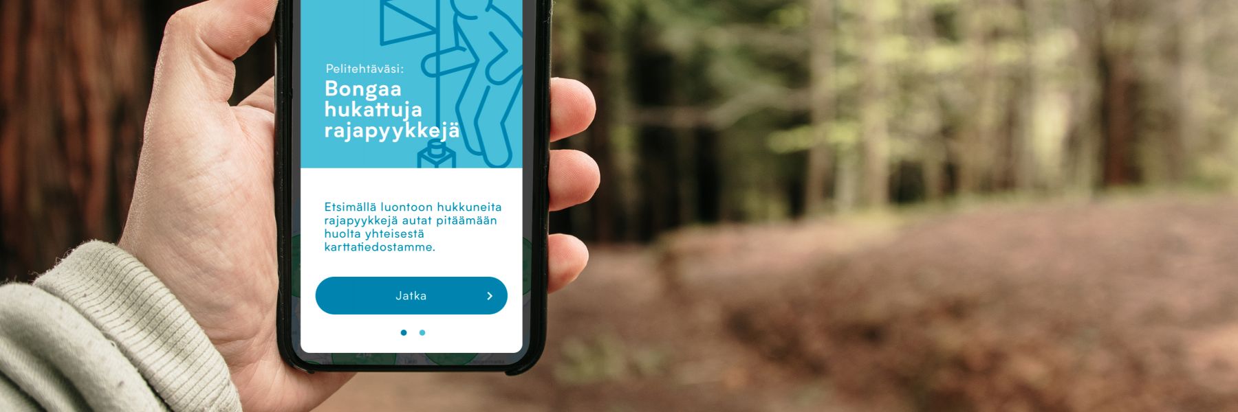 Metsämaisemassa käsi pitää kädessä kännykkää, jossa on auki Pyykkijahti-peli.