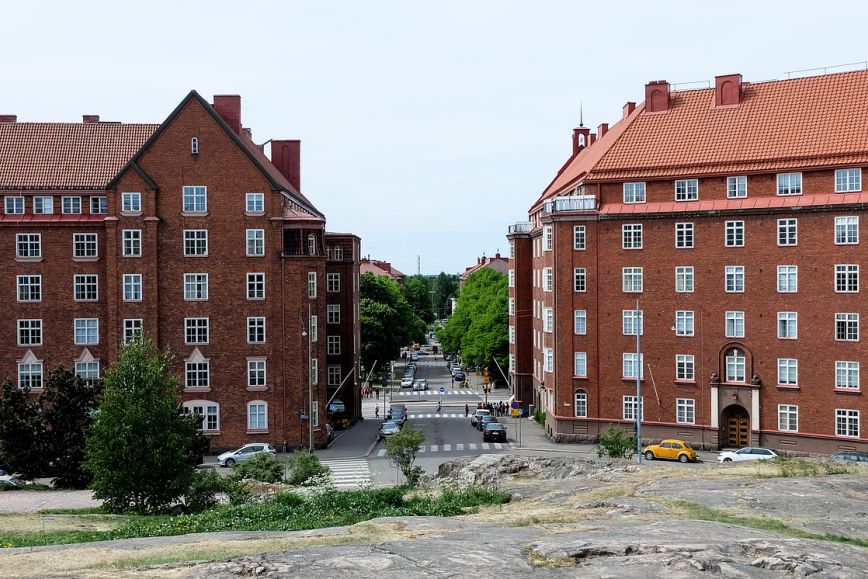 Kuvan etualalla näkyy kalliota. Keskellä kuvaa kulkee katu suoraan eteenpäin. Kadun molemmilla puolilla on oransseja tiilikerrostaloja. Kadun laidoilla on puisto.