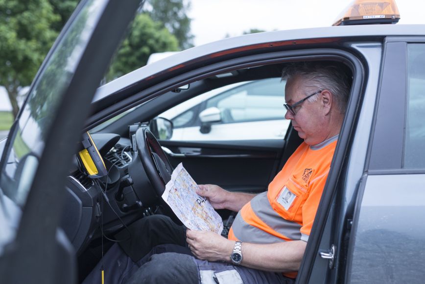 Kartoittaja istuu Maanmittauslaitoksen autossa ja katsoo paperista karttaa. Hänellä on oranssi työpaita yllään ja sen rinnassa henkilökortti.