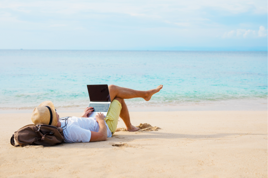 Mies työskentelee kannettavalla tietokoneella ja loikoilee rantahiekalla vaaleanturkoosin meren äärellä.