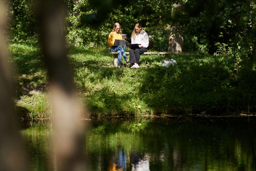 Två personer sitter på en bänk på en skogig strand med bärbara datorer i famnen.