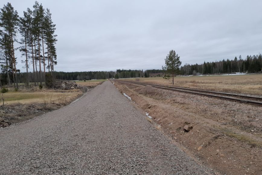 Kuvan keskellä näkyy eteenpäin kulkeva hiekkatie. Hiekkatien oikealla puolella on junarata. Molemmissa reunoissa näkyy peltoa ja puita.
