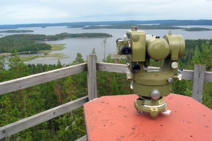 en teodolit, som är ett mätinstrument av metall, står uppställd på ett utsiktstorn i trä med ett insjölandskap i bakgrunden