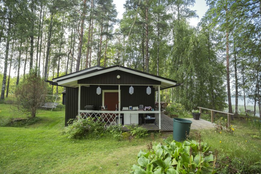 Pieni tummanruskea mökki järven rannalla vehreässä maisemassa.