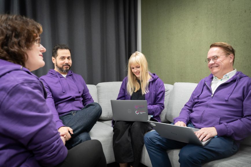 Neljä henkilöä violetit hupparit päällä sohvalla työskentelemässä kannettavilla tietokoneilla.