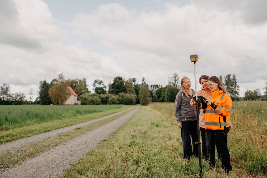 Tien reunassa seisoo kolme naista rinnakkain. Yhdellä naisista on yllään oranssi huomiotakki.  Hän pitelee teknistä laitetta, joka muodostuu korkeasta kepistä ja ruudusta, jota naiset katselevat. Vehreä maalaismaisema, kesä. Taustalla peltoja ja talorakennus.