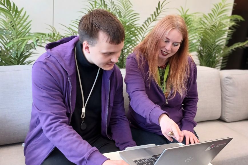 Kaksi henkilöä istuu harmaalla sohvalla ja työskentelee yhdessä kannettavan tietokoneen äärellä. Molemmilla on violetit hupparit, ja toinen osoittaa sormella tietokoneen näyttöä. Taustalla on vihreitä kasveja.