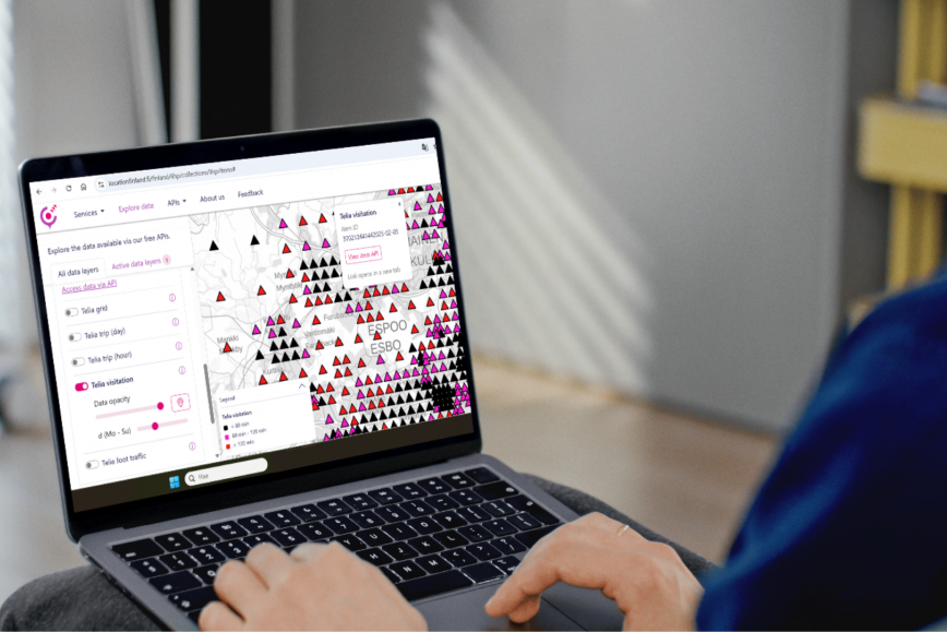 Henkilö käyttää kannettavaa tietokonetta, jonka näytöllä on värikäs karttasovellus. Kartassa näkyy mustia ja pinkkejä kolmioita ruudukossa sekä paikkakuntien nimiä, kuten Espoo. Vasemmalla puolella on valikkoja ja asetuksia.