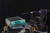 På bilden finns FGI:s rymdgoniospektrometerutrustning. Den klara punkten i mitten är ett prov.  Linsen på armen samlar in ljuset i fibern och för det till den gröna spektrometern i förgrunden.