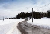 Vinterlandskap med väg och snötäckta åkrar. Längre bort syns skog och ett hus.