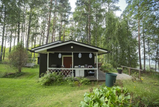 A small dark brown cottage on the shore of a lake in a lush landscape.