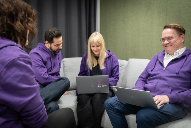 Neljä ihmistä istuu sohvalla, kahdella on kannettava tietokone. Kaikilla ihmisillä on päällään violetit hupparit.