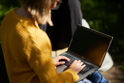 Keltapaitainen työntekijä, jolla on vaalea polkkatukka ja silmälasit, työskentelee ulkona puistossa kannettavalla tietokoneella, jossa on tietosuojakalvo.