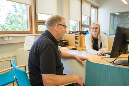 En medelålders kund med glasögon sitter vid en servicedisk. På andra sidan disken sitter en leende kundrådgivare framför en dator. Hen har glasögon och ljust, långt hår.
