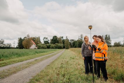 Tre kvinnor står bredvid varandra vid kanten av en väg. En av kvinnorna har en orange varseljacka på sig. Hon håller i en teknisk apparat som består av en hög stav och en ruta som kvinnorna tittar på. Grönt landsbygdslandskap, sommar. Åkrar och en husbyggnad i bakgrunden.