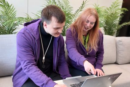 Två personer sitter på en grå soffa och arbetar tillsammans vid en bärbar dator. Båda har lila huvtröjor, och den ena pekar på datorskärmen. Gröna växter syns i bakgrunden.