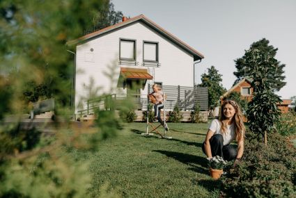Tontilla on uusi valkoinen omakotitalo. Pihanurmikolla lapsi hyppää estettä keppihevosella ja etualalla aikuinen tekee puutarhan istutustöitä iloisen näköisenä.